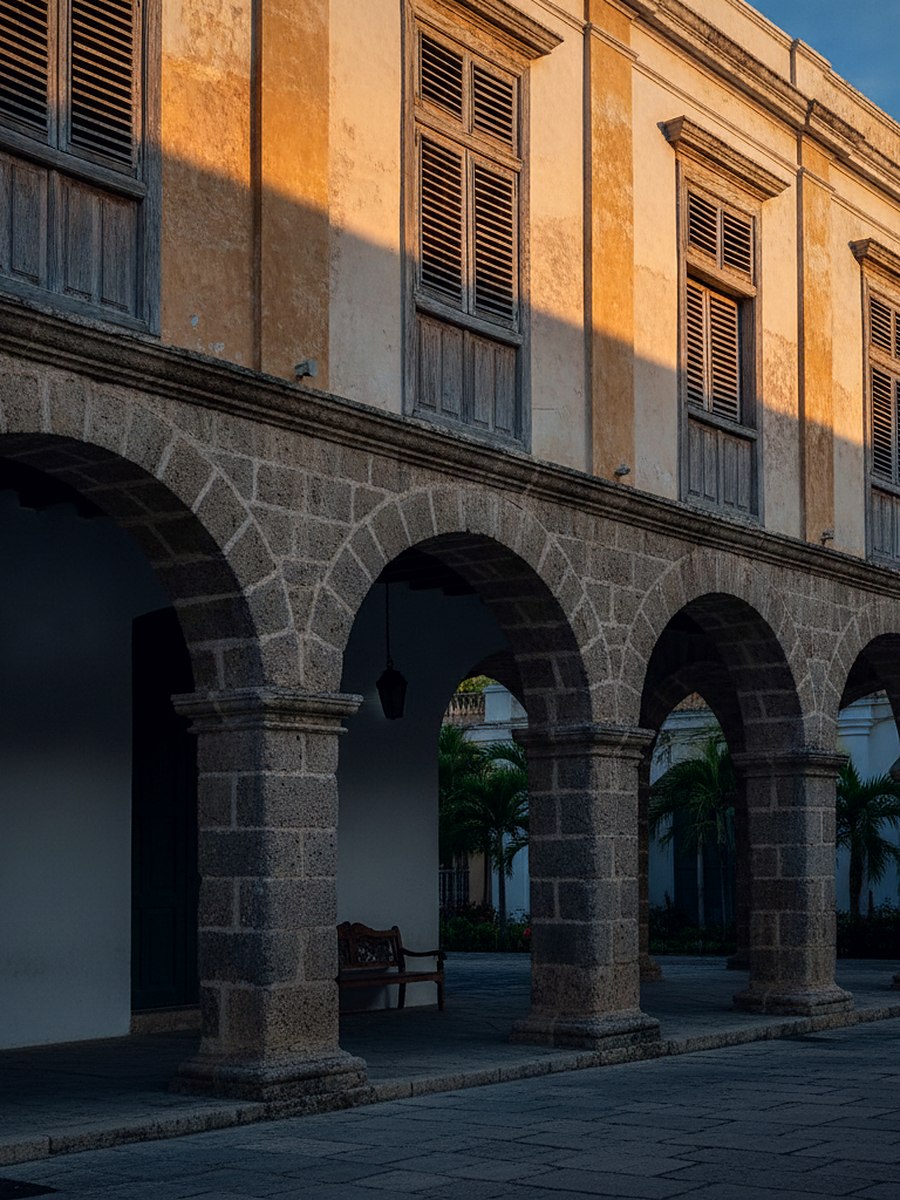 A tall pastel colonial facade with iron window grilles and deep shadow under an arcade